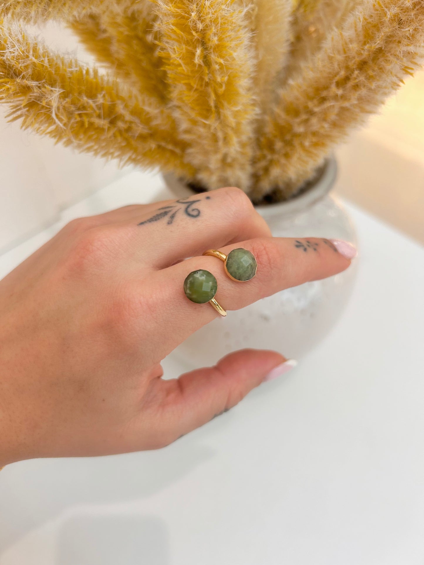 A close-up of a hand wearing an adjustable gold ring with round green jade stone tops, posed in front of a white vase with dried golden grass on a white surface.