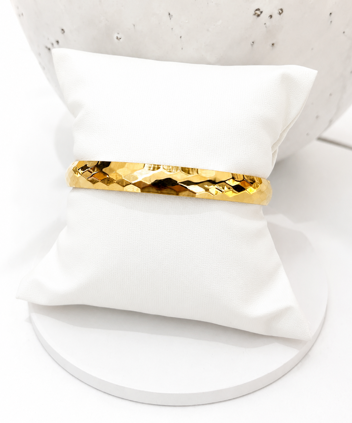 A gold geometric textured bangle bracelet displayed on a white fabric jewelry pillow, set against a white background with a white speckled ceramic bowl.