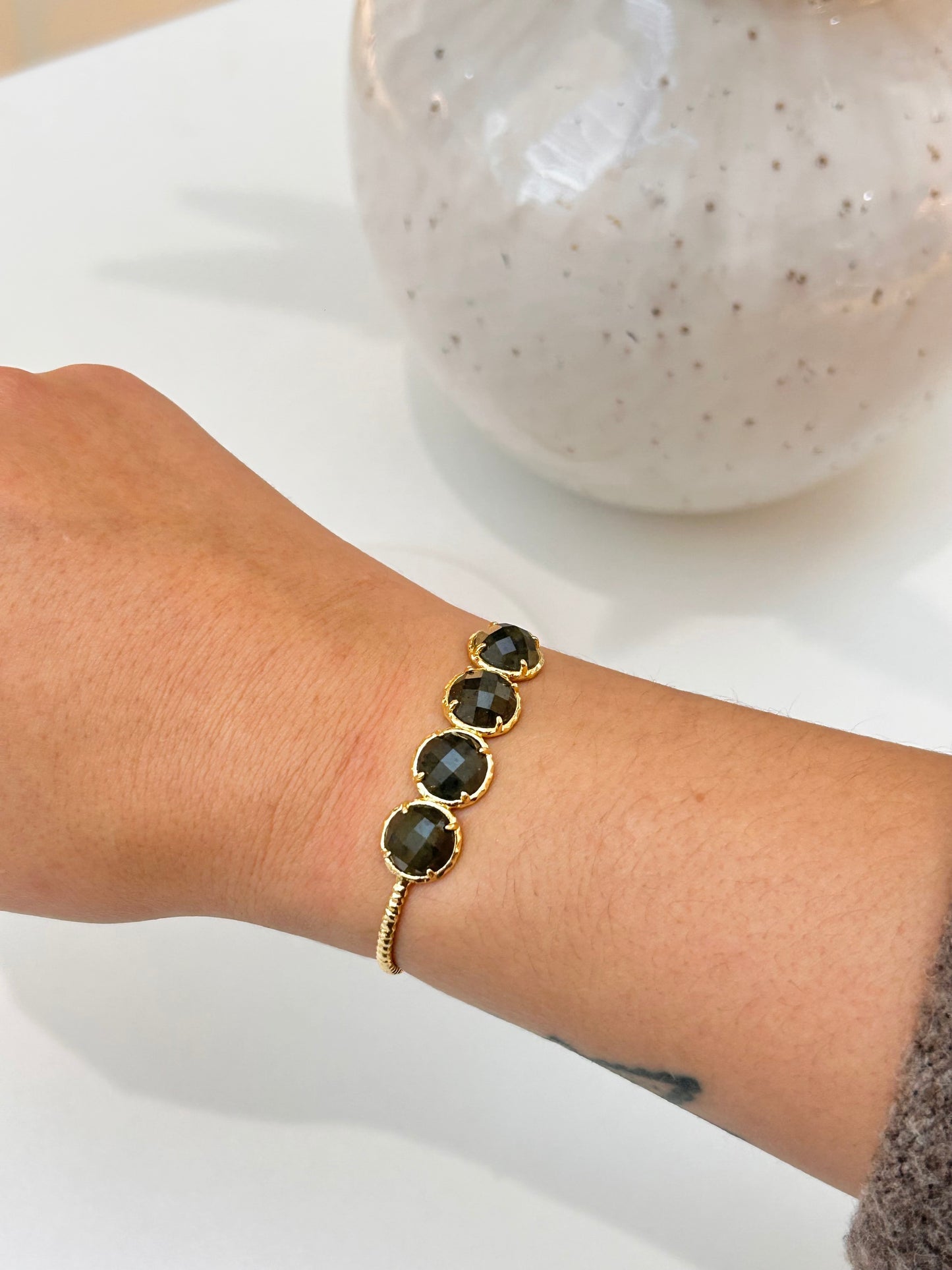 A gold-toned bracelet with three round Labradorite stones, worn on a person's wrist against a white background with a blurred ceramic vase.