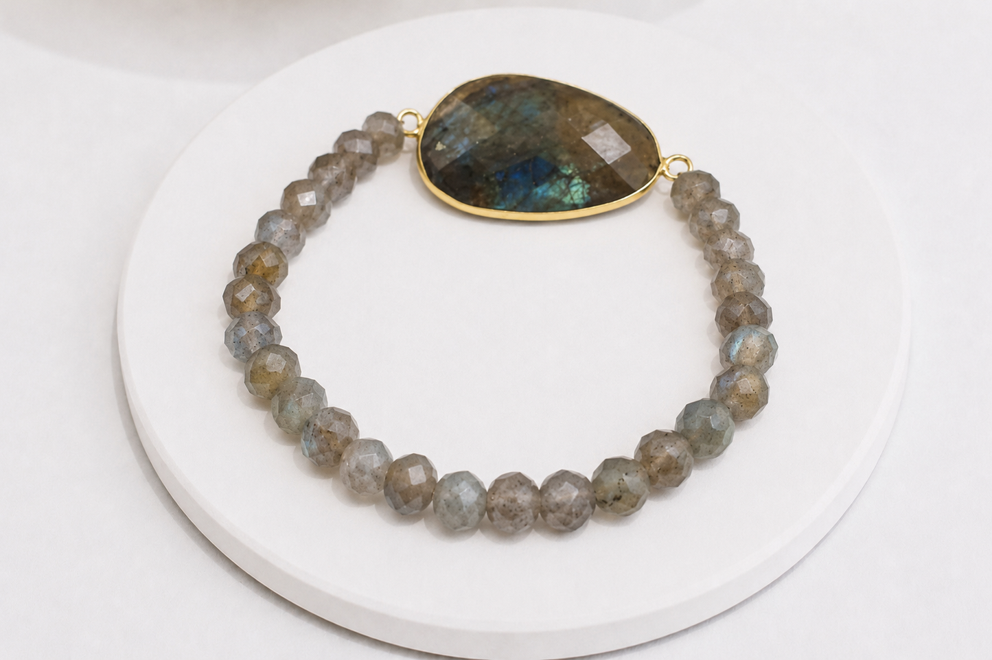 A beaded stretch bracelet made of faceted gray and brown labradorite beads, featuring a large teardrop-shaped natural labradorite stone clasp with a gold-tone border, displayed on a white circular stand.