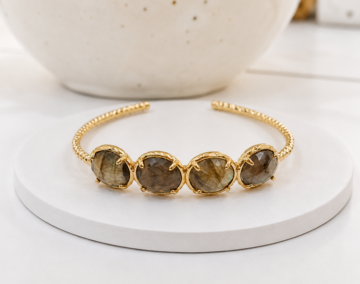 A gold cuff bracelet featuring four oval labradorite gemstones, displayed on a white round pedestal with a ceramic bowl in the background.
