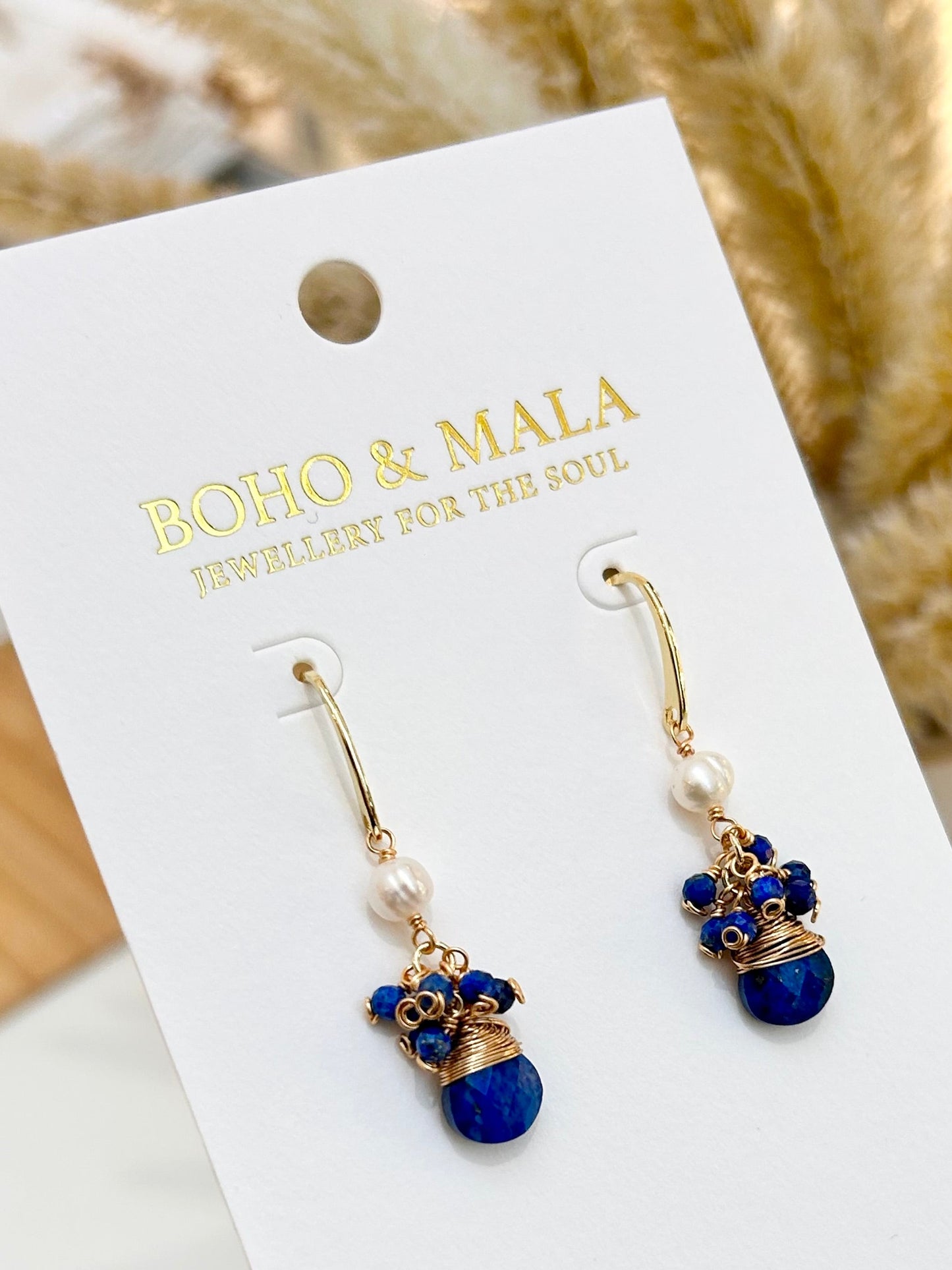 
                  
                    Pair of blue and gold earrings on a BOHO & MALA branded card held by a hand.
                  
                