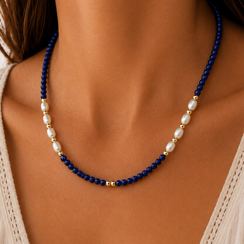 A close-up of a woman wearing a lapis lazuli beaded necklace with deep blue beads, white freshwater pearls, and small gold spacer beads, paired with a cream-colored lace-trim top.