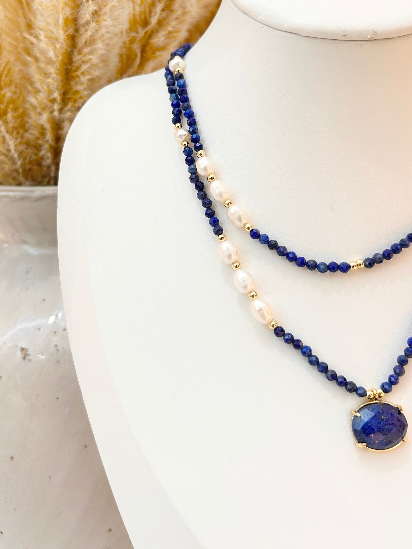 A double-strand necklace displayed on a white mannequin bust, featuring deep blue lapis lazuli beads, white freshwater pearls, and gold-tone metal accents with a round blue stone pendant.