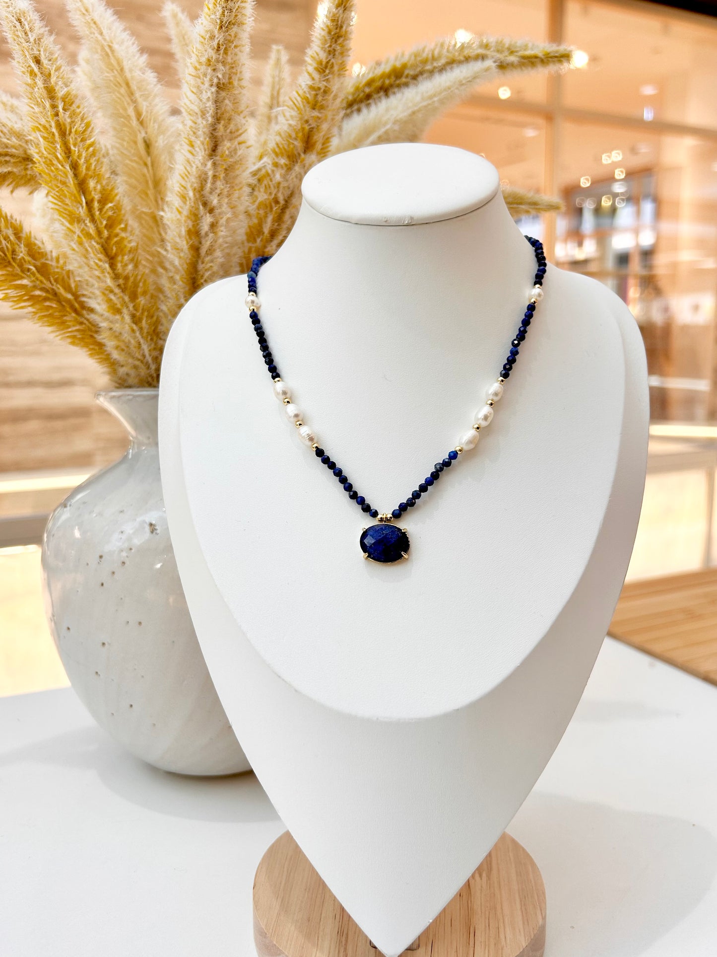 A beaded necklace with lapis lazuli and white freshwater pearl and a lapis lazuli oval pendant, displayed on a white jewellry bust with dried grass in the background.
