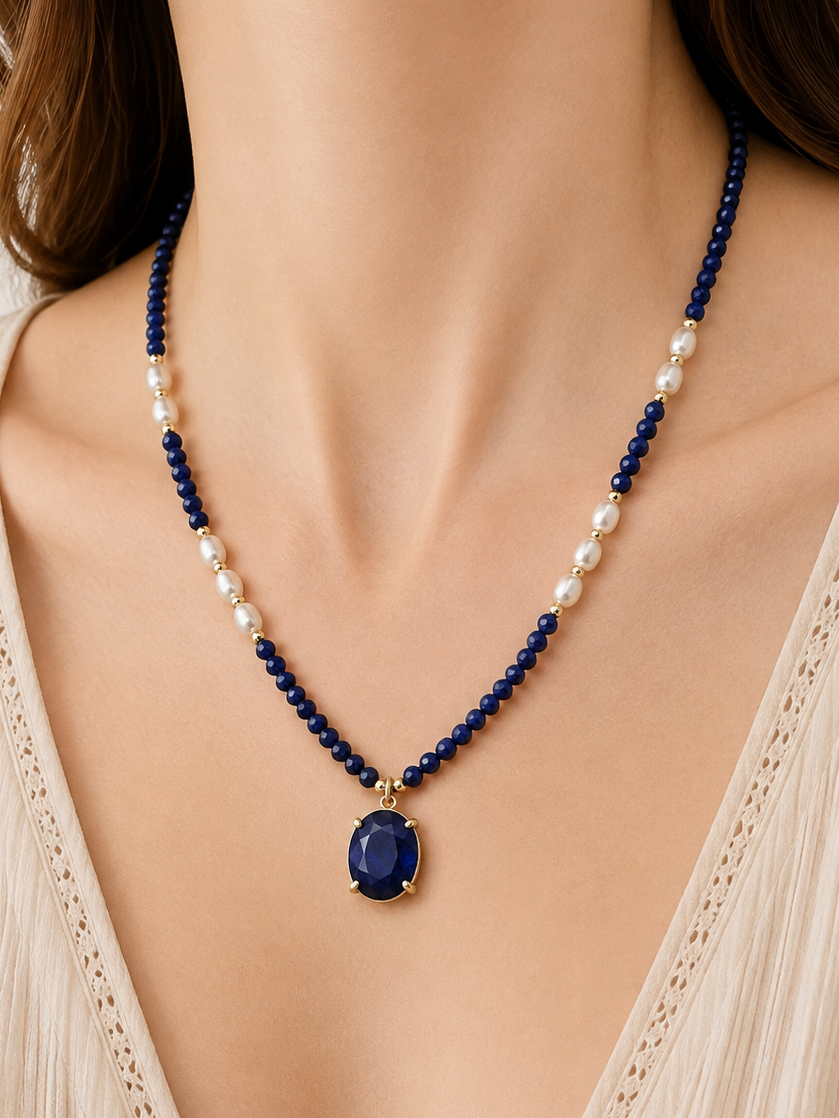 A close-up of a woman wearing a beaded necklace with lapis lazuli beads, white pearl accents, and a central oval blue stone pendant in a gold-tone setting.
