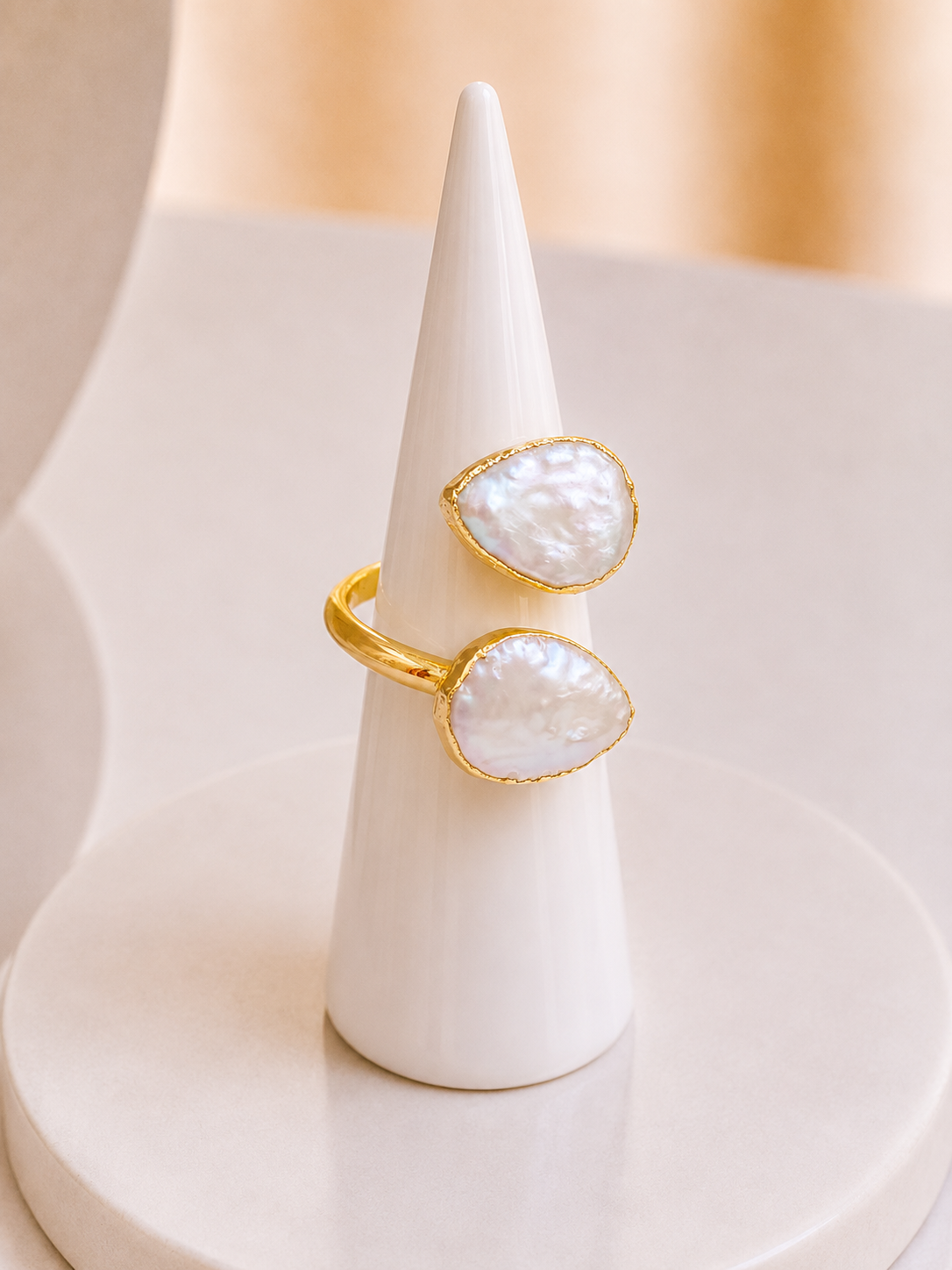 Two gold teardrop-shaped white mother-of-pearl stones on an adjustable ring displayed on a white cone jewelry stand against a soft beige background.