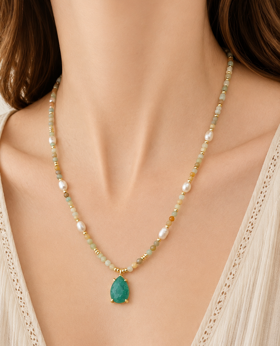 A close-up of a woman wearing a delicate beaded necklace with a amazonite teardrop pendant. The necklace features small white pearls, light green beads, and gold-toned details, styled with a cream-colored lace-trim top.