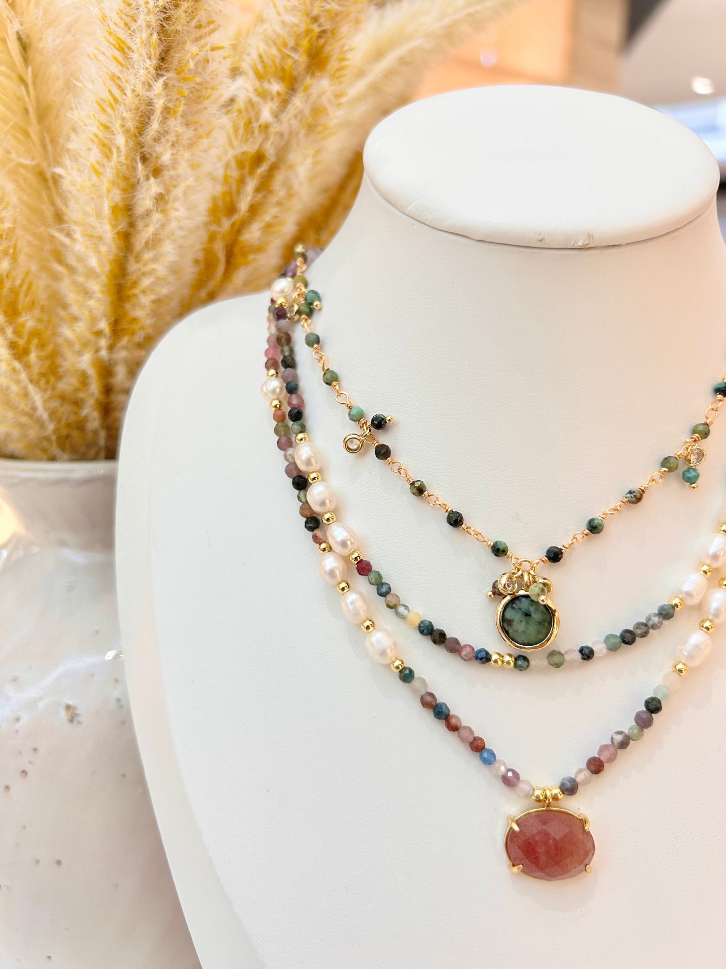 A multi-strand beaded necklace displayed on a white jewelry bust. The necklace features layers of white pearls, small multicolored natural beads, gold-toned spacer beads, and two central pendants: a green oval stone and a larger dusty rose oval stone, both accented with gold-tone detailing.