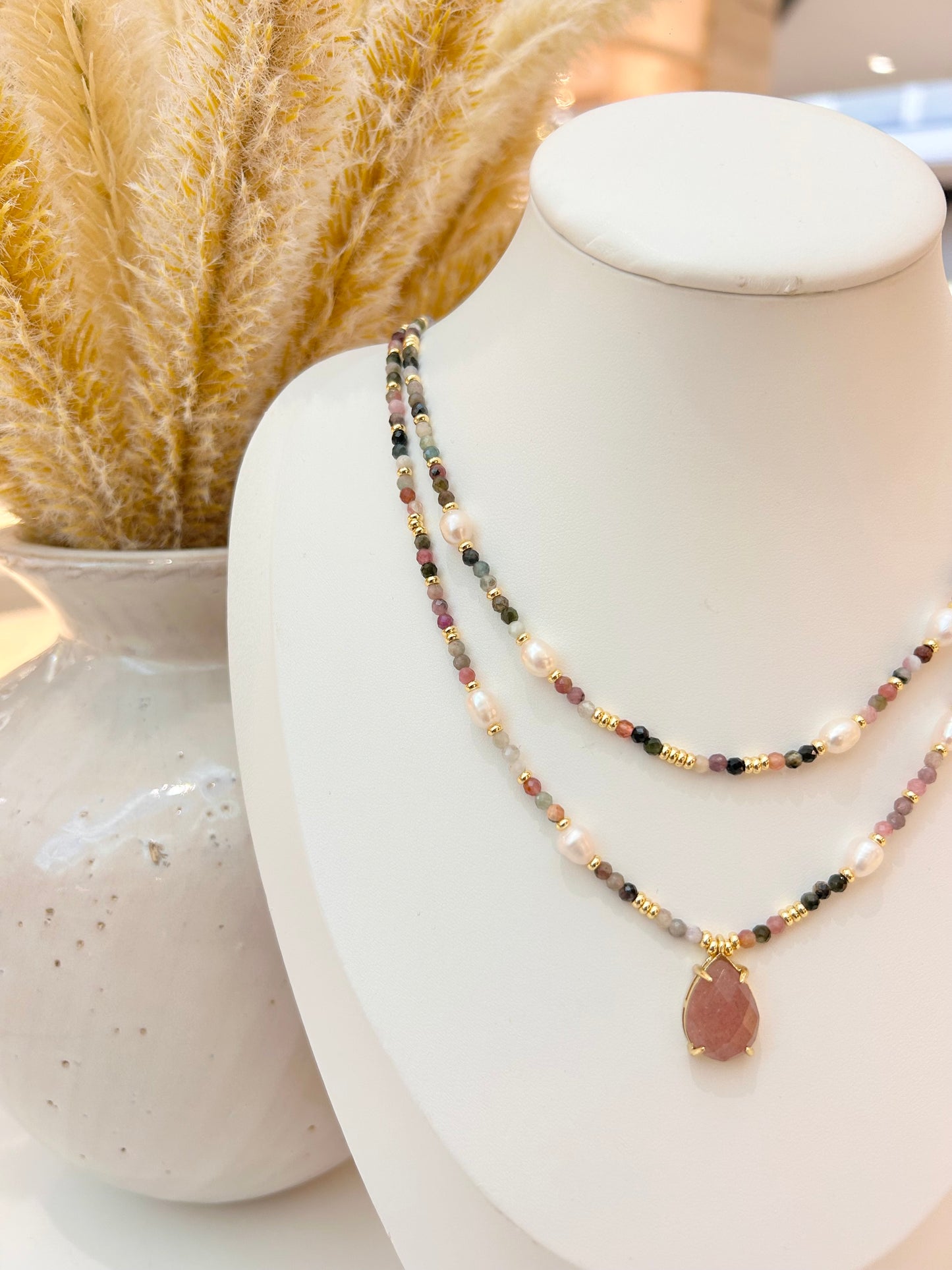 A double-strand beaded necklace with a teardrop-shaped sunstone pendant, displayed on a white jewellery bust next to a vase with dried pampas grass.