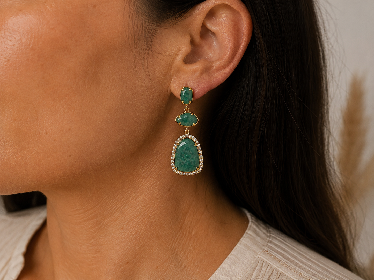 A close-up of a woman wearing a gold drop earring with three green amazonite stones and a halo of small clear stones.