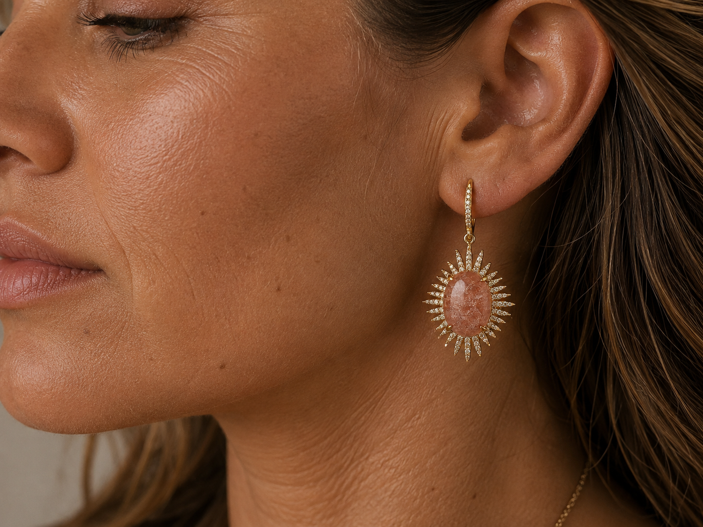 A close-up side profile of a woman wearing a gold sunburst drop earring with a pink stone, set against her ear and neck.