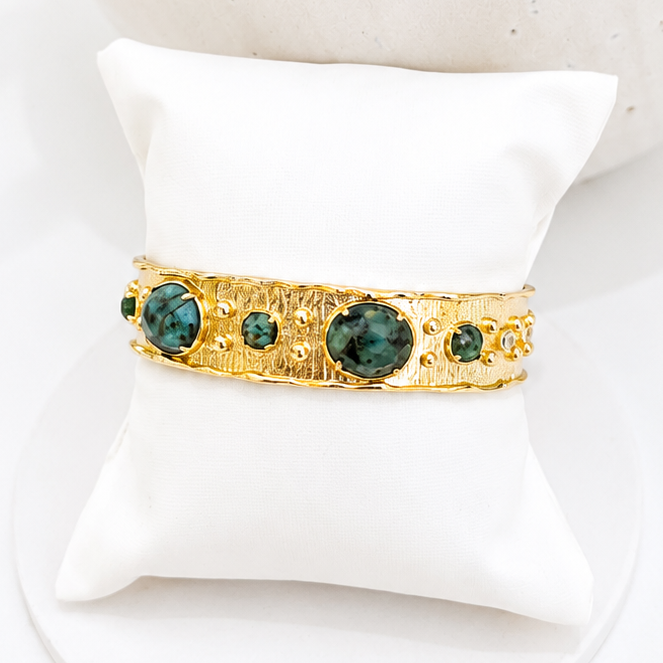 
                  
                    A gold cuff bracelet with  African Turquoise accents, displayed on a white fabric jewellery pillow.
                  
                