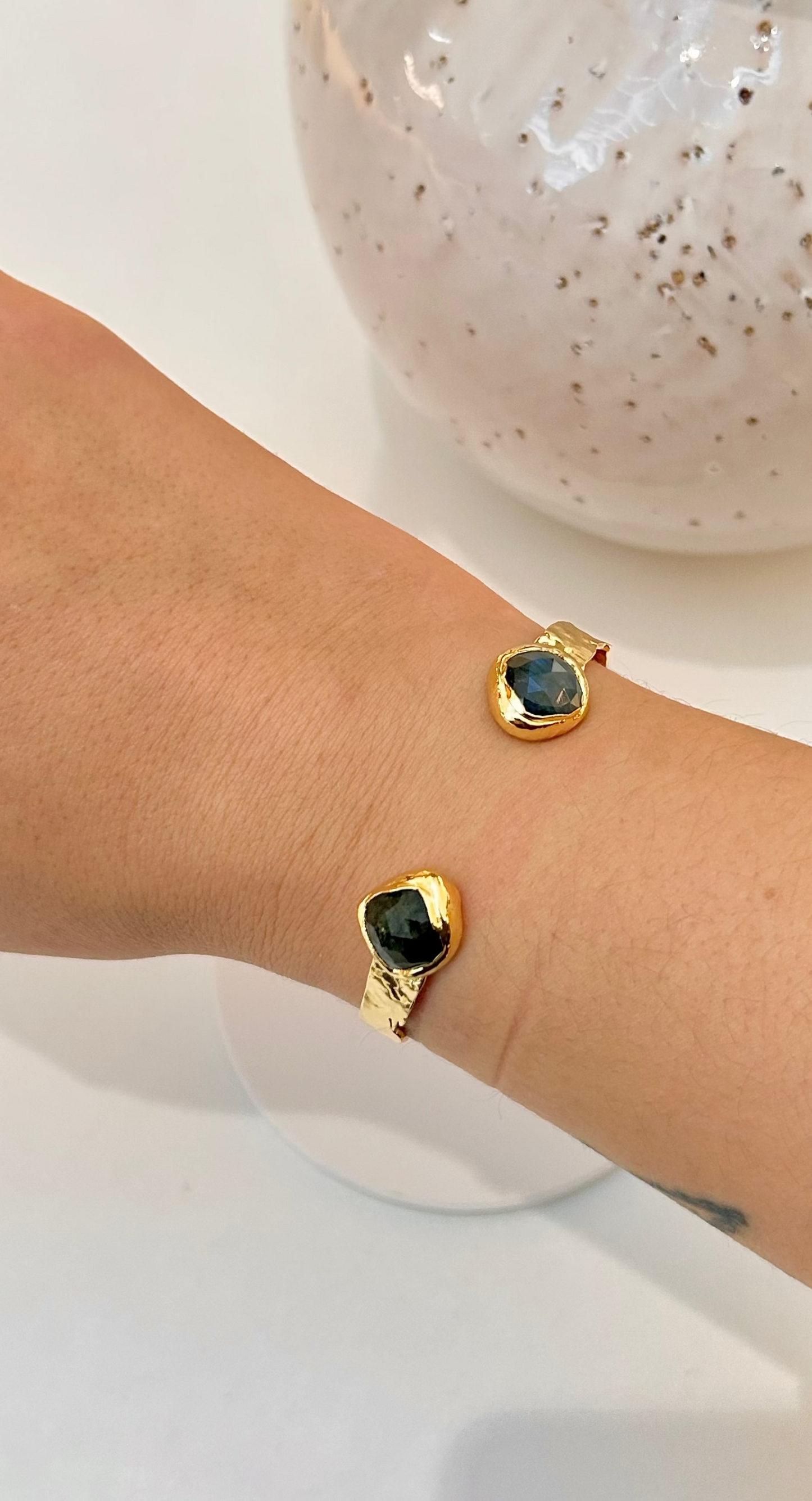 A close-up of a person's wrist wearing gold cuff bangle with labradorite stones, set against a white tabletop with a speckled ceramic vase in the background.