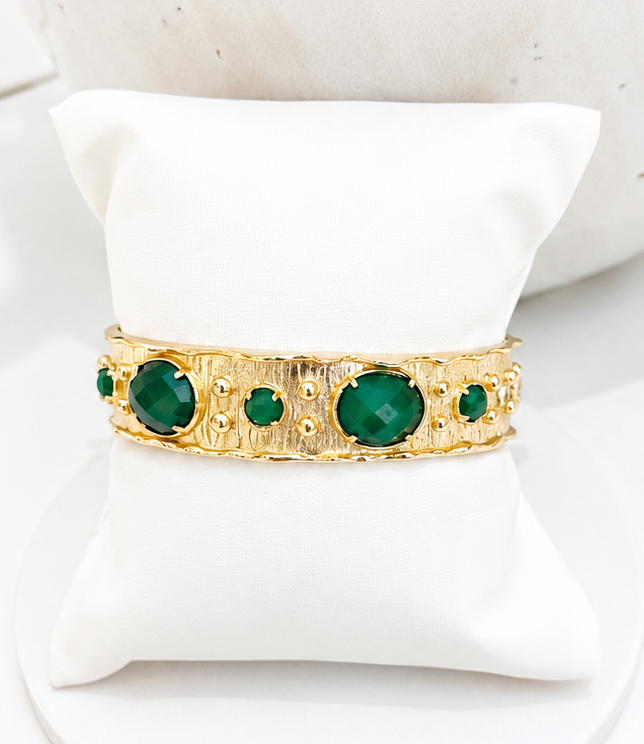 
                  
                    A gold bangle bracelet with large and small green green gemstones, displayed on a white cushion.
                  
                