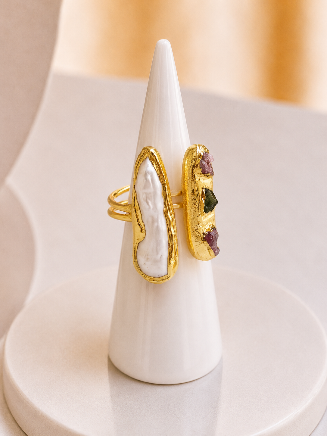 A gold ring displayed on a white cone stand, featuring a large white textured stone on one side and a smaller side panel with freshwater pearl, purple and green stones on a gold base.