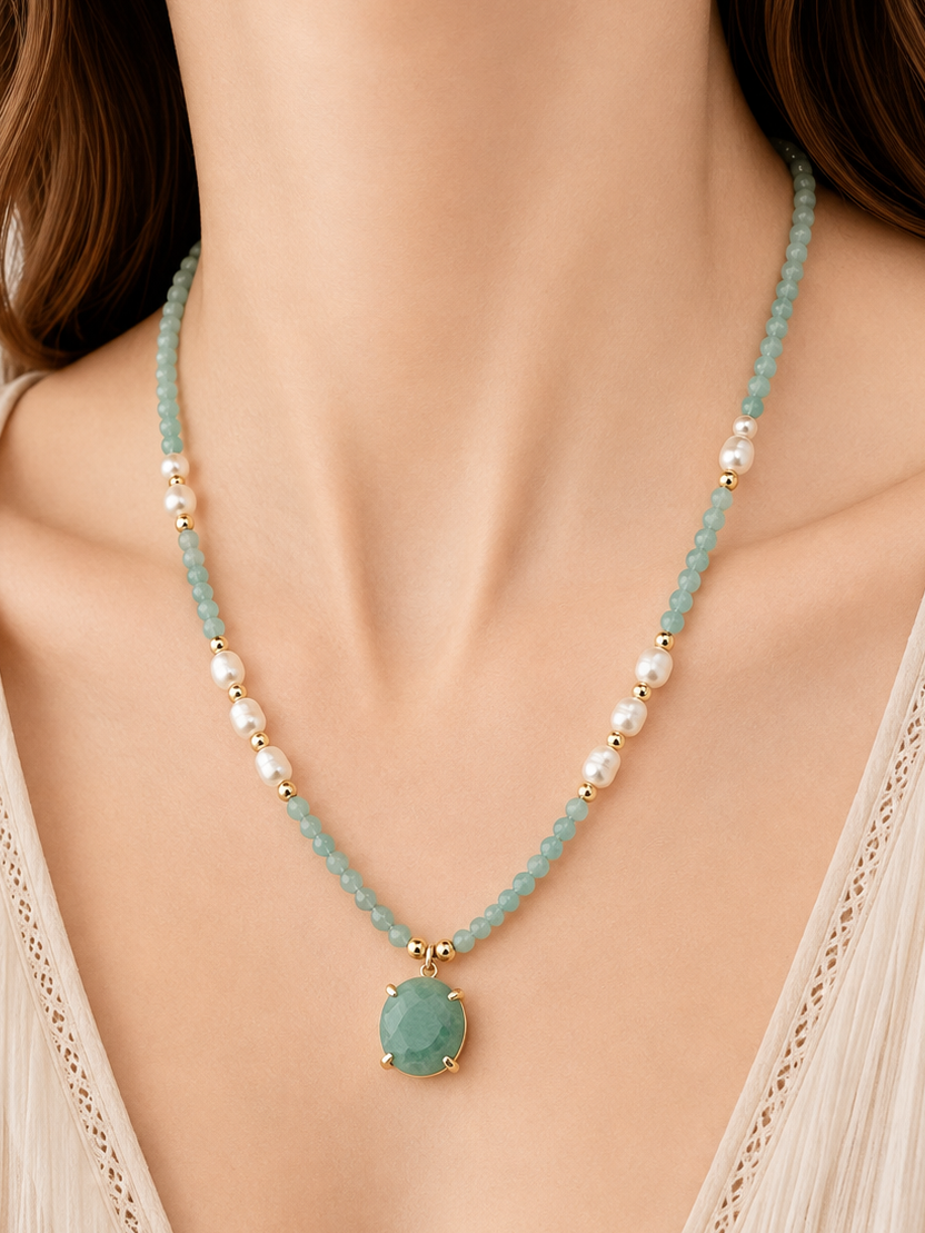 A close-up of a woman wearing a delicate necklace with amazonite light blue-green beads, white pearl-like beads, and a central oval green stone pendant with gold-tone detailing.