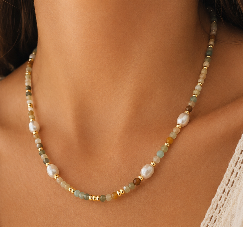 A close-up of a woman wearing a delicate beaded necklace with white pearls and small mixed amazonite stones paired with a cream-colored lace-trimmed top.