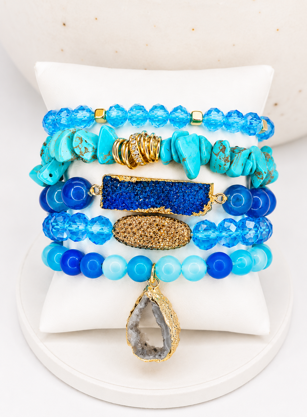 A stack of five beaded bracelets displayed on a white jewelry stand. The bracelets feature various shades of blue beads, turquoise-colored stone pieces, a rectangular blue crystal charm, a textured gold oval charm, and a teardrop-shaped clear crystal pendant with a gold frame.