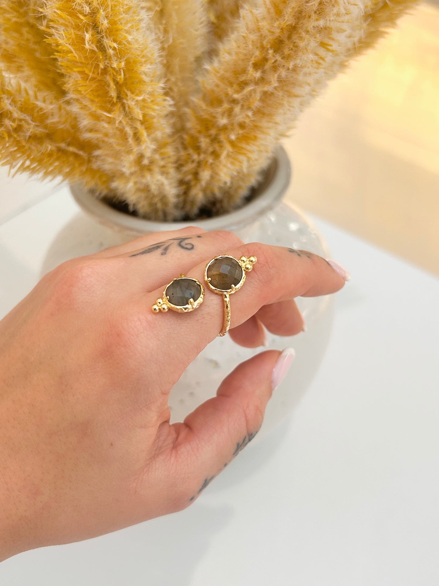 A close-up of a hand wearing an adjustable gold ring with oval labradorite stones, posed in front of a vase of dried pampas grass on a white surface.