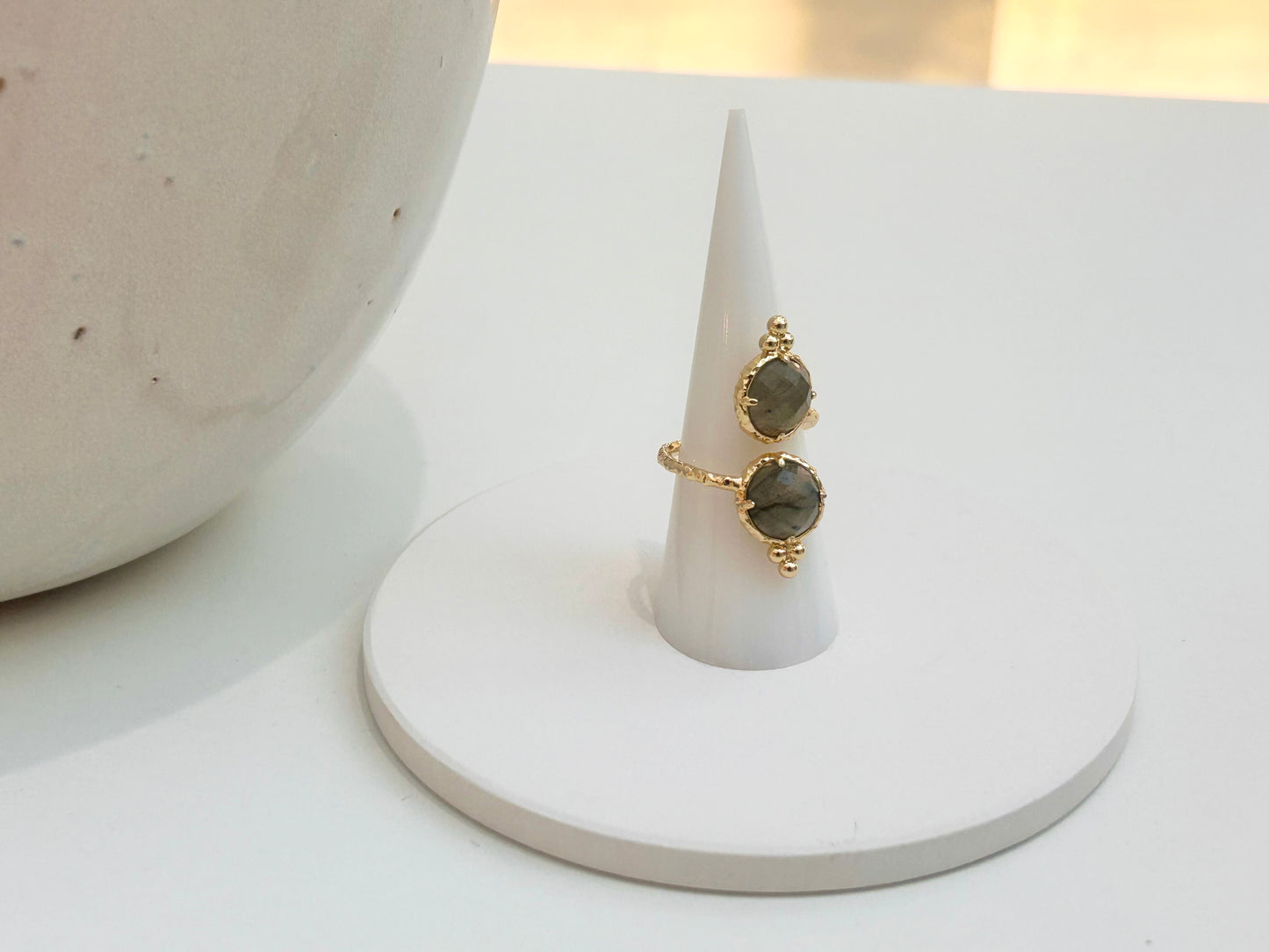 
                  
                    A gold ring with adjustable stacked oval labradorite stones displayed on a white cone ring holder, set against a clean white background with a ceramic vessel partially visible on the left.
                  
                