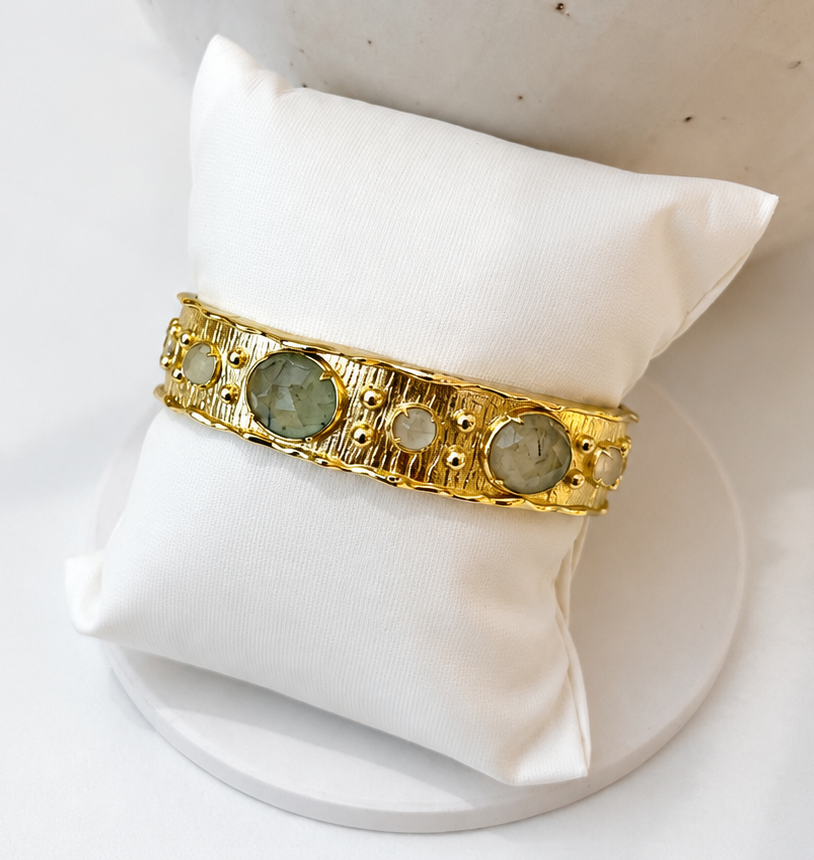 
                  
                    A tetured gold bangle bracelet with three round prehnite gemstones and small clear stones, displayed on a white fabric jewelry pillow.
                  
                