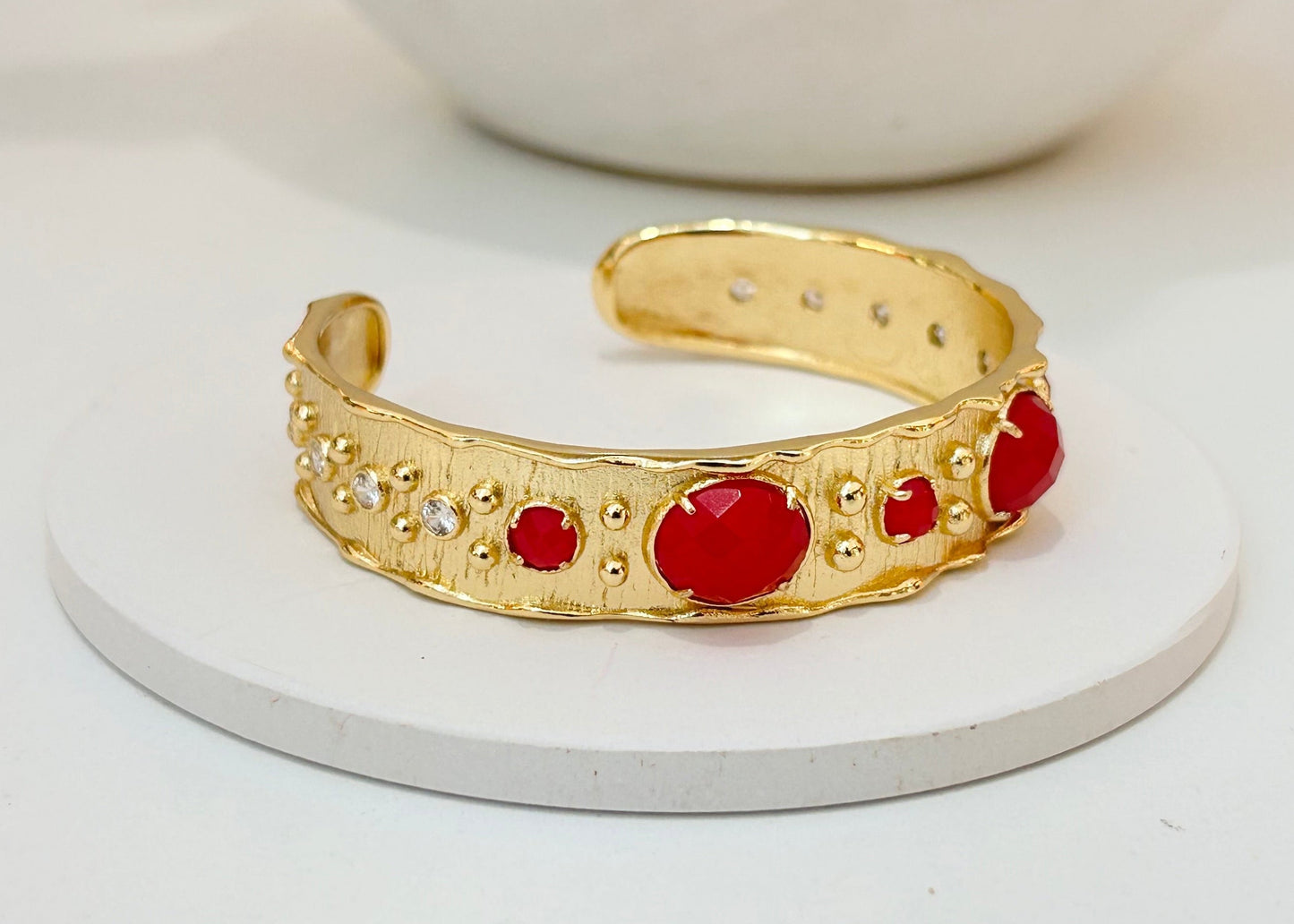 
                  
                    A gold cuff bracelet with red jade gemstones and small clear rhinestones, displayed on a round white coaster.
                  
                