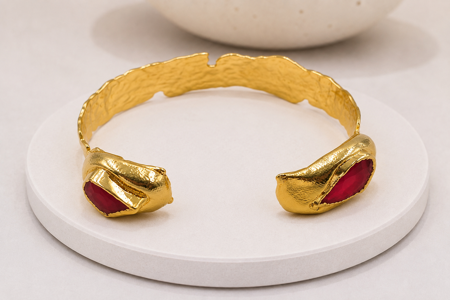 A hammered gold cuff bracelet with two sculptural, organic-shaped gold ends set with deep red inlays, red coral, displayed on a round white ceramic riser.