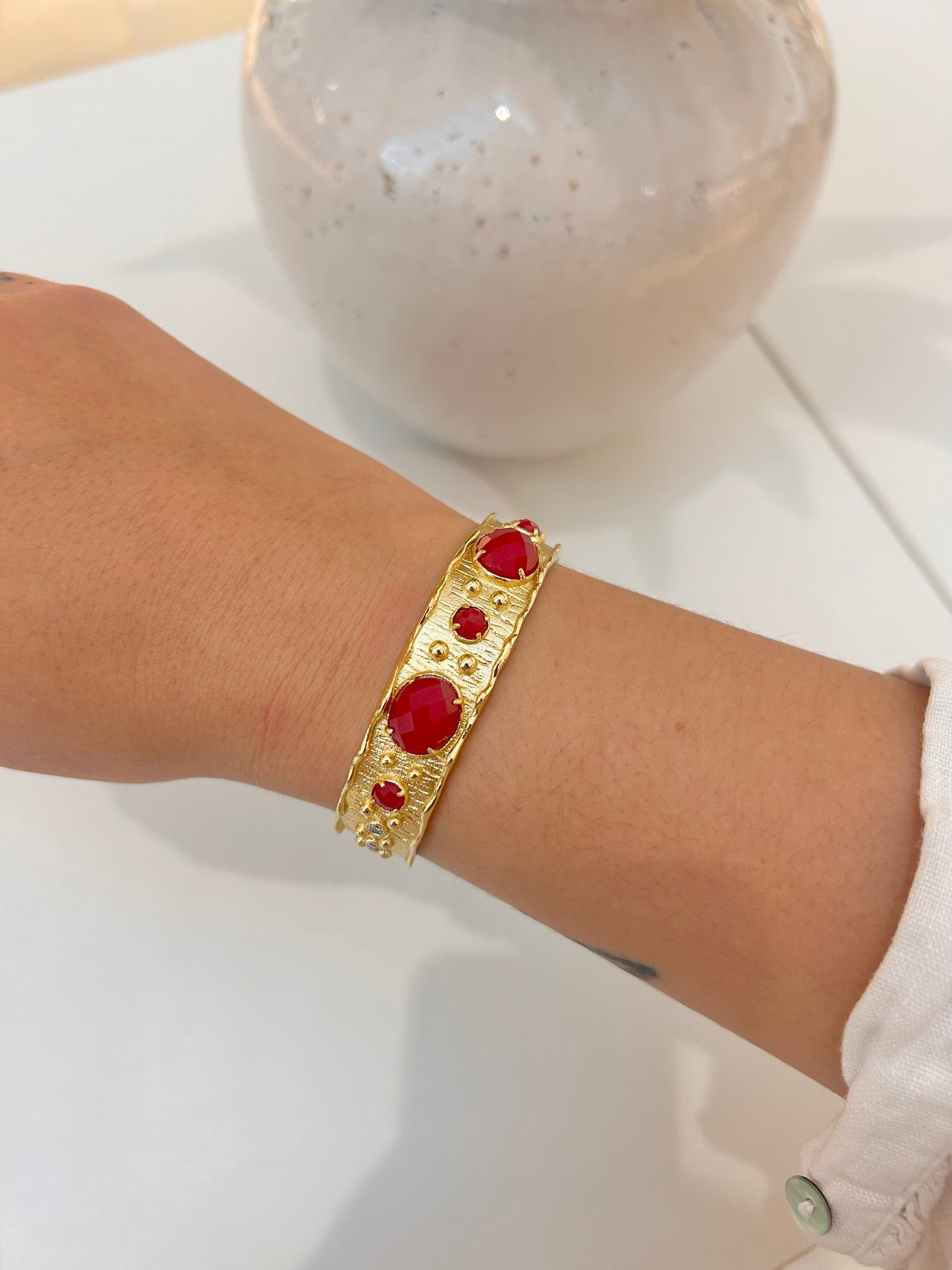 A gold bangle bracelet with red jade gemstone accents worn on a person's wrist, shown against a white background with a ceramic vase in the soft-focus background.