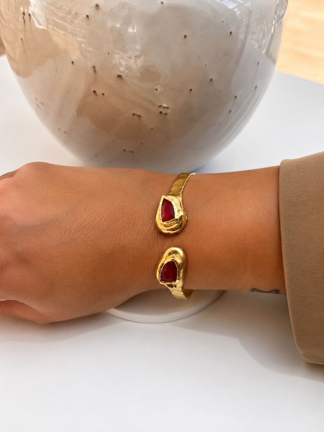 A gold cuff bracelet with two teardrop-shaped red jade gemstone inlays, worn on a person's wrist with a beige sleeve visible.