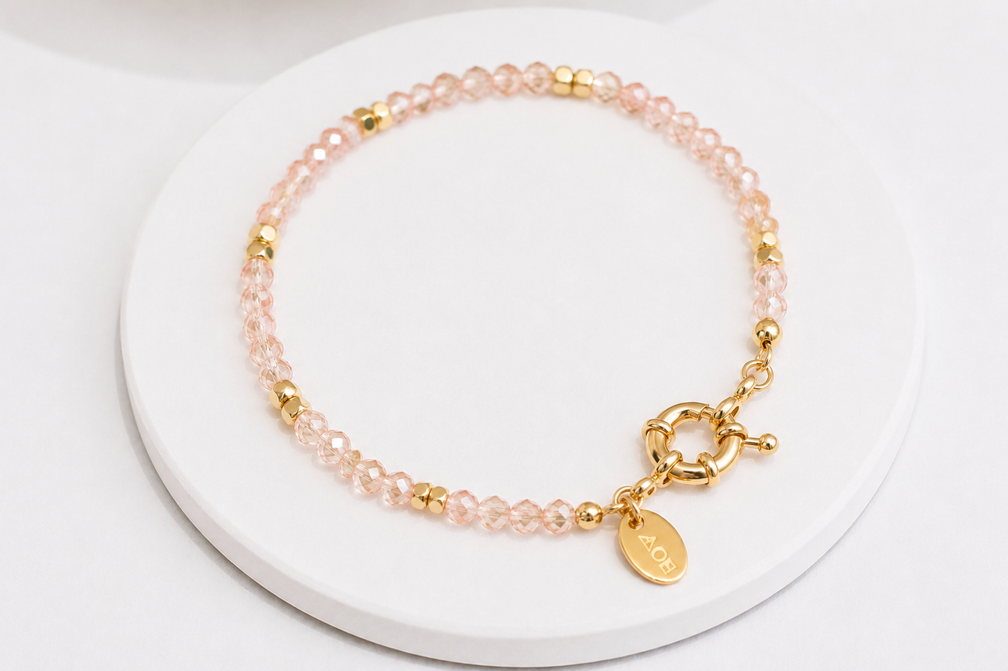 A delicate beaded bracelet with rose quartz beads, small gold spacer beads, and a gold toggle clasp with a round gold tag, displayed on a white circular stand.