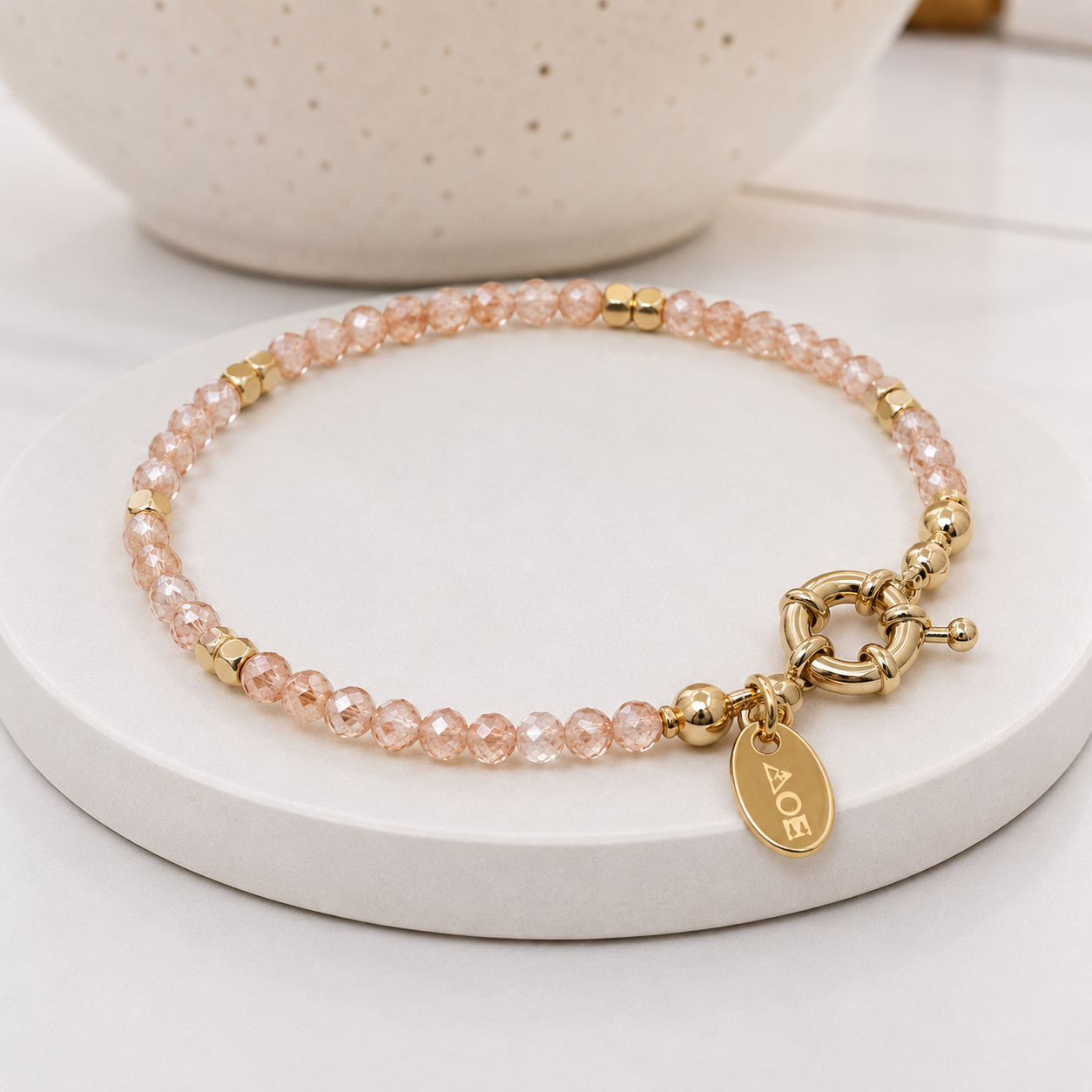 
                  
                    A delicate beaded bracelet with rose quartz beads and gold-toned metal accents, including a toggle clasp and a round tag. The bracelet is displayed on a round white ceramic stand with a soft-focus beige bowl in the background.
                  
                