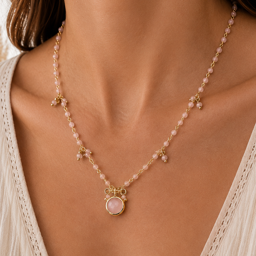A close-up of a model wearing a delicate gold and pink beaded necklace with a central oval pink rose quartz stone pendant, styled with a cream-colored lace top.
