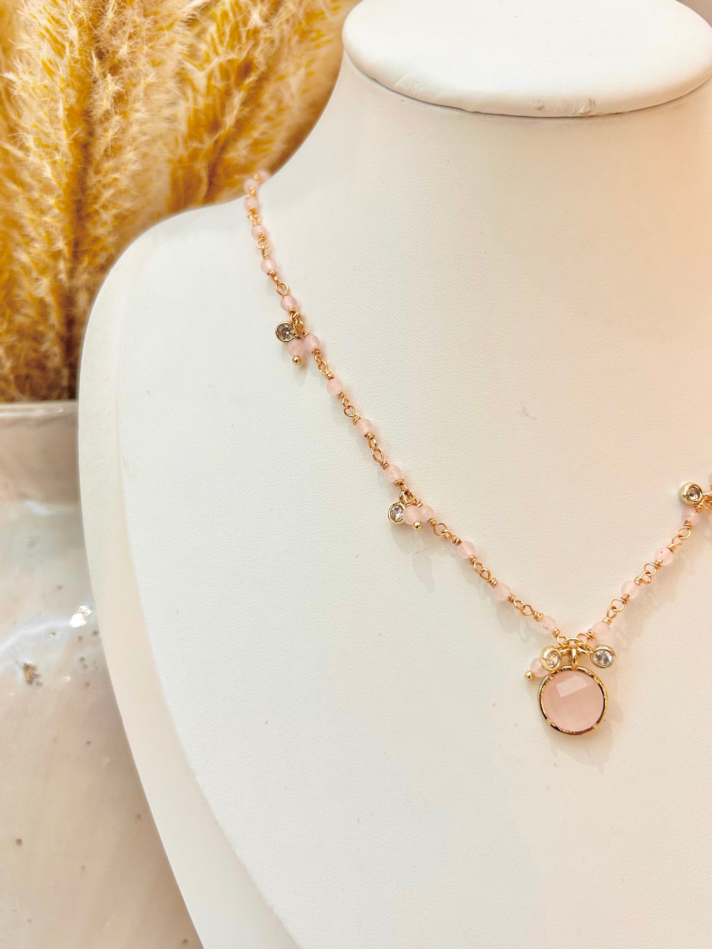 A delicate necklace displayed on a white jewelry bust, featuring a gold-tone chain with small pale pink rose quartz  beads, tiny cubic zirconia charms, and a central round pink rose quartz  stone pendant.