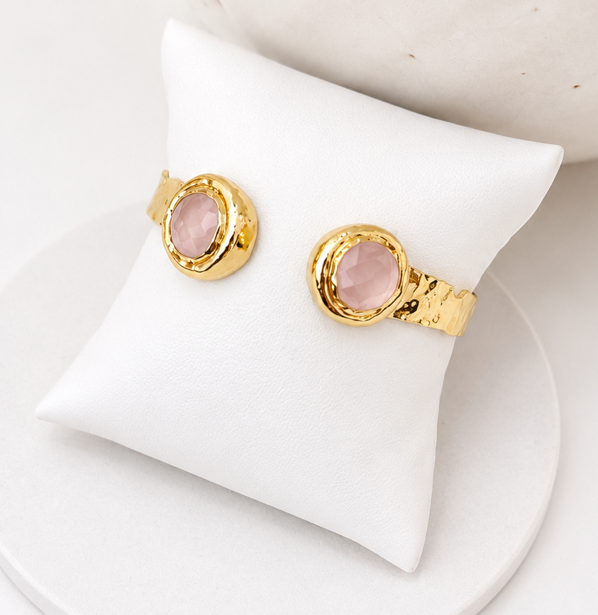 
                  
                    A pair of gold-toned rings with oval rose quartz gemstones, displayed on a white cushion.
                  
                