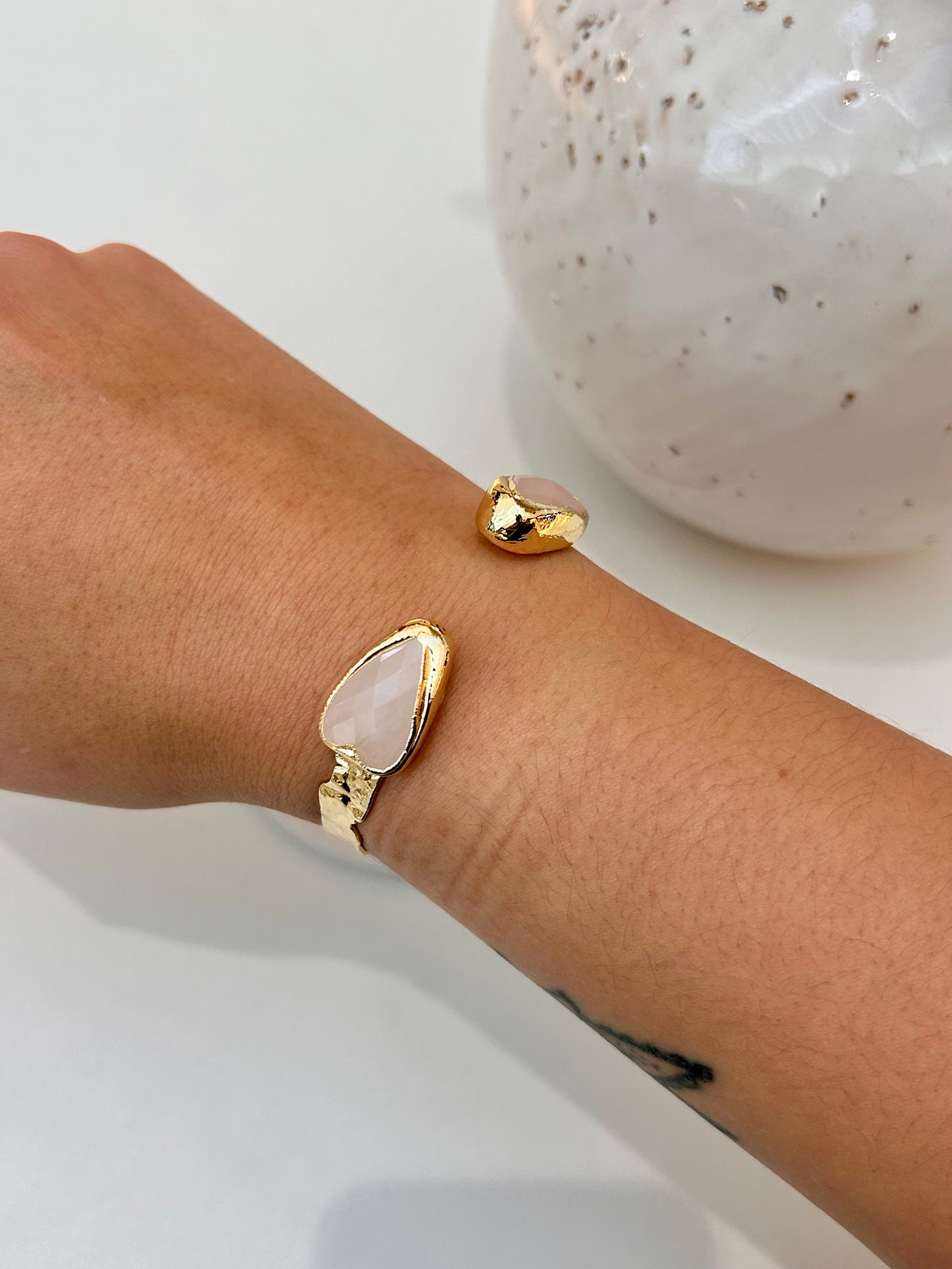 A gold-tone cuff bracelet with a teardrop-shaped pale pink rose quartz stone, shown worn on a person's wrist next to a matching small gold charm, with a white speckled ceramic vase in the background.