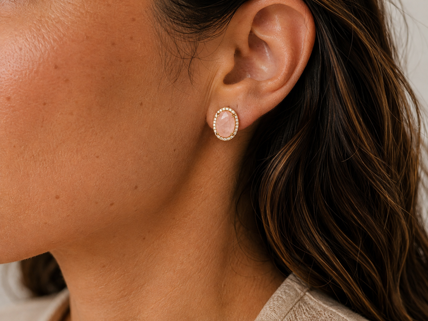 A close-up of a model's ear wearing an oval stud earring with a pink rose quartz stone surrounded by a gold and white stone border.
