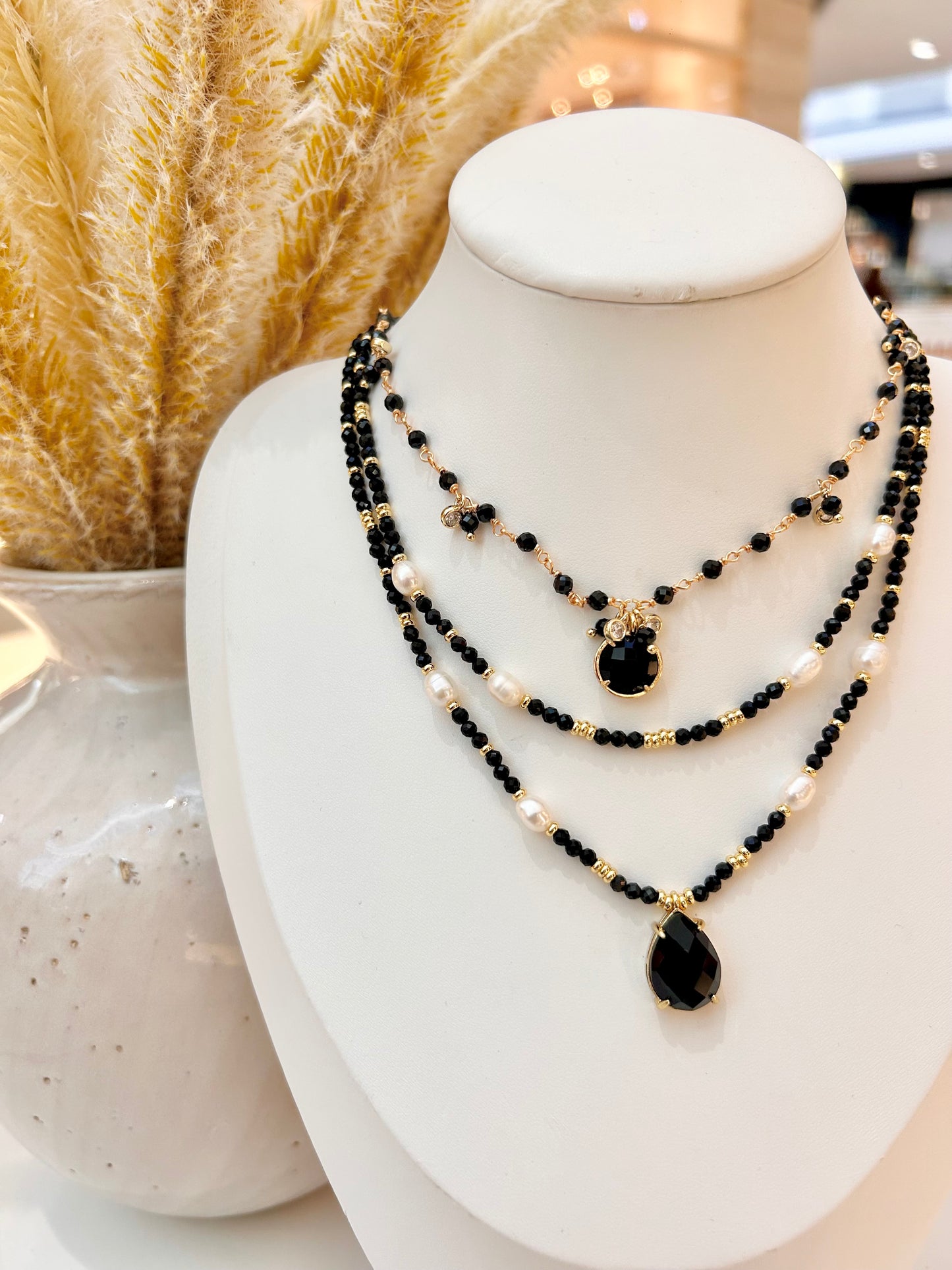 A three-layer beaded necklace displayed on a white mannequin bust. The necklace features layers of black obsidian beads, white freshwater pearls, and small gold spacer beads, culminating in a large black teardrop pendant and a smaller black oval pendant.