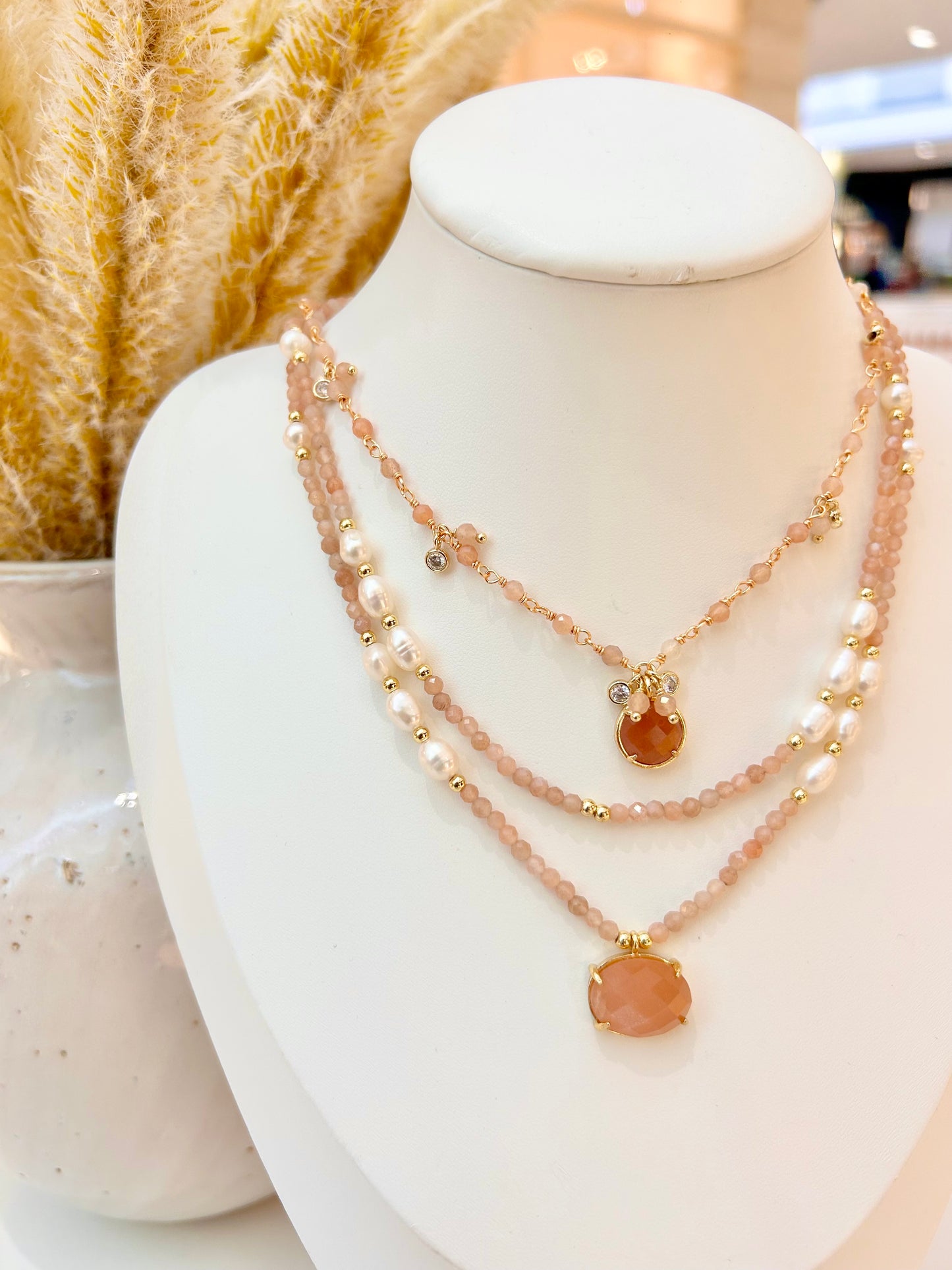A three-layered sunstone and pearl necklace with gold-toned accents and a sunstone pendant, displayed on a white jewelry bust.
