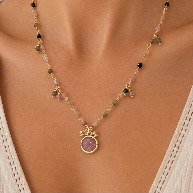 A close-up of a model wearing a delicate gold-toned beaded necklace with a round strawberry quartz stone pendant, styled with a cream-colored lace-trimmed top.