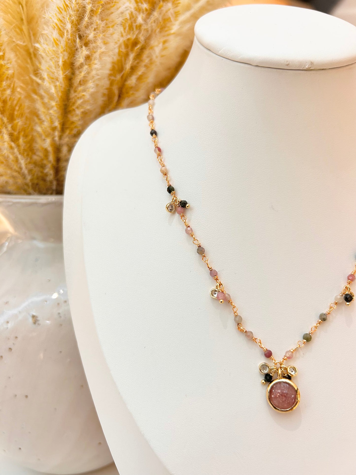 
                  
                    A delicate gold-tone beaded necklace with a round strawberry quartz stone pendant, displayed on a white jewellery bust.
                  
                