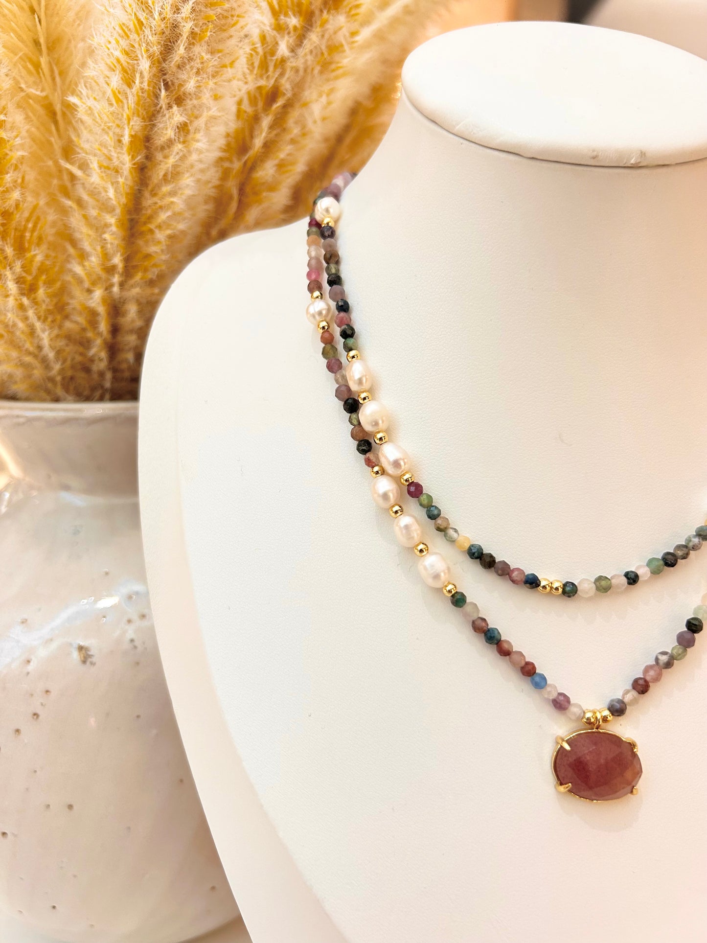 A double-strand beaded necklace displayed on a white mannequin bust. One strand features white pearls, while the other is made of small multicolored beads with gold spacer accents and a round maroon pendant in a gold bezel.