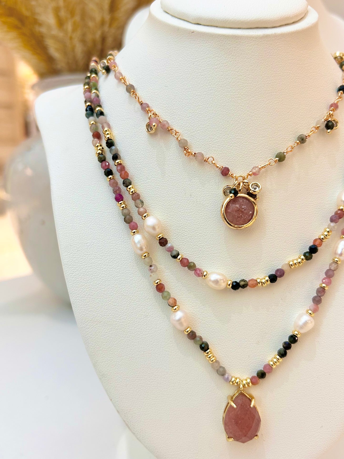 A multi-strand beaded necklace displayed on a white jewellery bust. The necklace features gold-tone chains, white pearl-like beads, faceted beads in pink, green, and black, and two teardrop-shaped pink stone pendants in gold-tone settings.