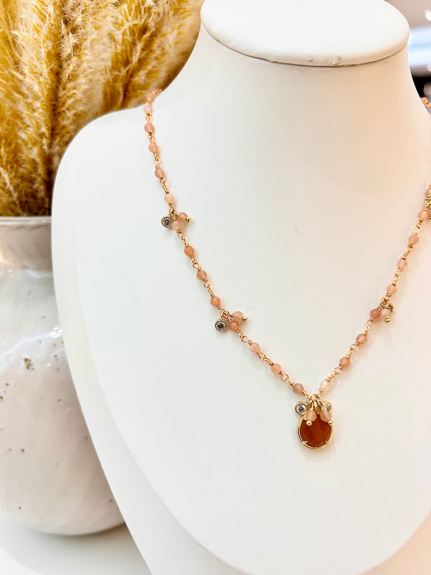 A delicate necklace displayed on a white jewelry bust. The necklace features a rose gold chain with small peach-colored beads, tiny cubic zirconia stones, and a central round sunstone pendant with a gold-toned setting.
