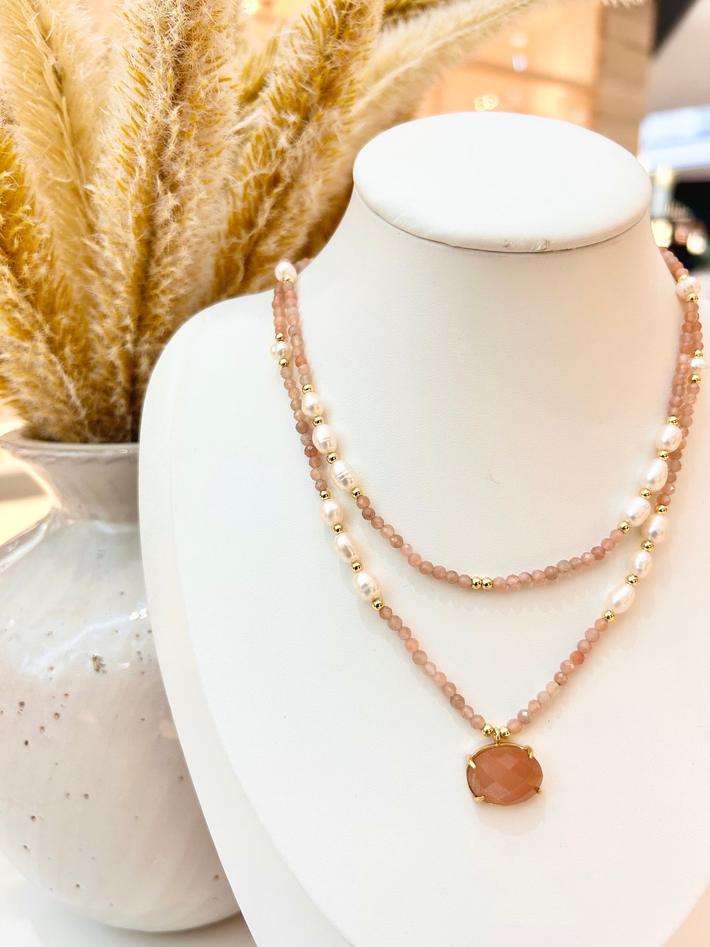 A double-strand necklace displayed on a white jewellery bust. The necklace features sunstone faceted beads, white pearl beads, and a central peach-pink oval pendant with gold-tone settings.