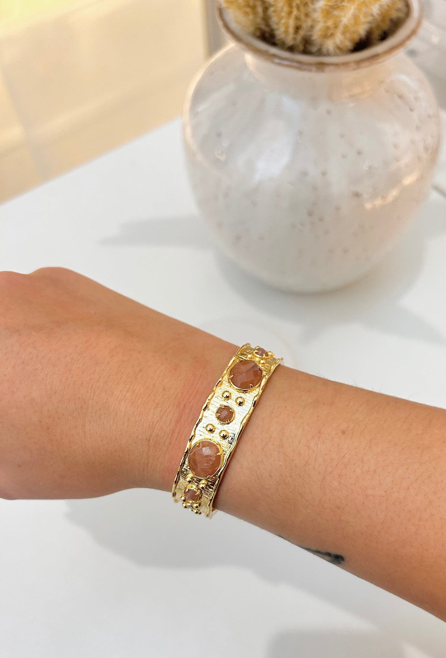 A gold-toned cuff bracelet with round sunstone accents worn on a person's wrist, shown against a white tabletop with a blurred ceramic vase in the background.