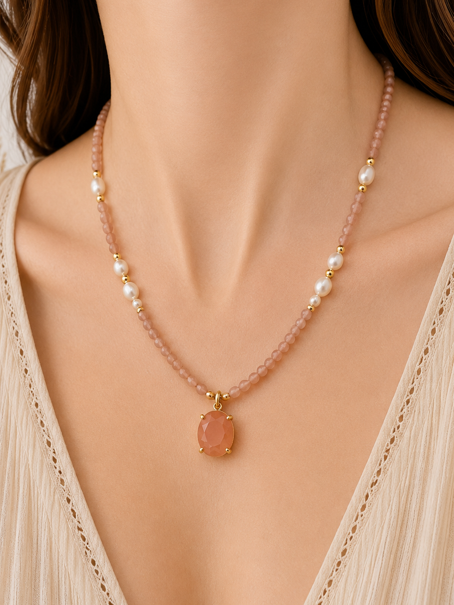 A close-up of a model wearing a delicate sunstone beaded necklace with white freshwater pearl beads and a pink oval pendant in gold-tone settings, paired with a cream-colored pleated top.