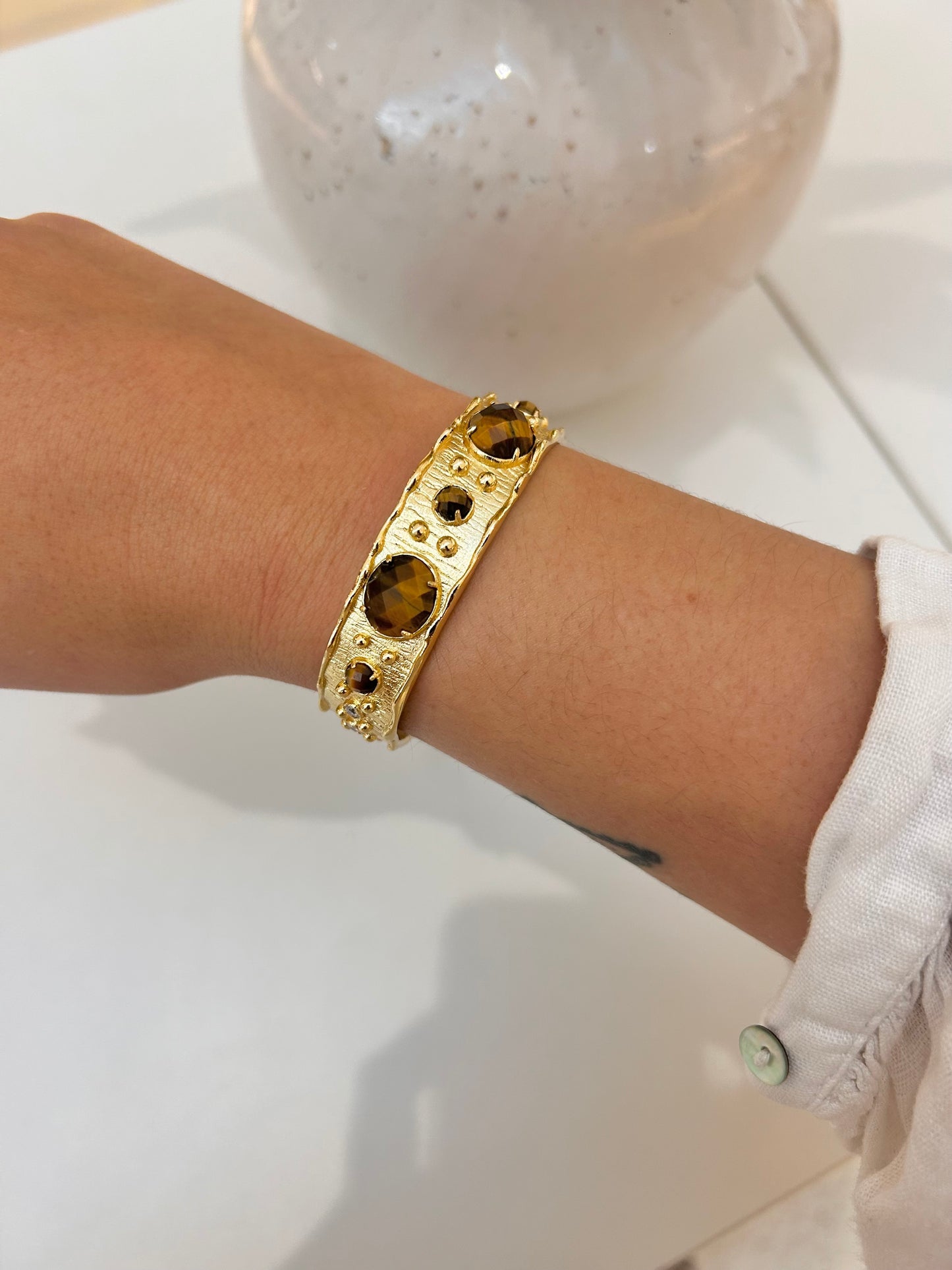 A gold-toned cuff bracelet with large tigers eye stones and small dark accents, worn on a person's wrist against a white tabletop background.