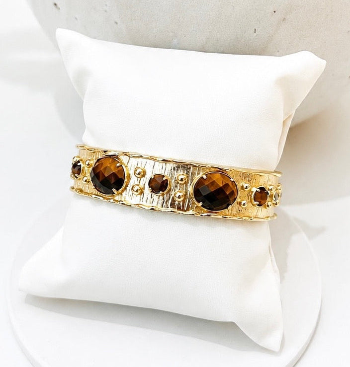 
                  
                    A gold-toned cuff bracelet with large tigers eye  gemstones and small gold beads, displayed on a white fabric jewellery pillow.
                  
                