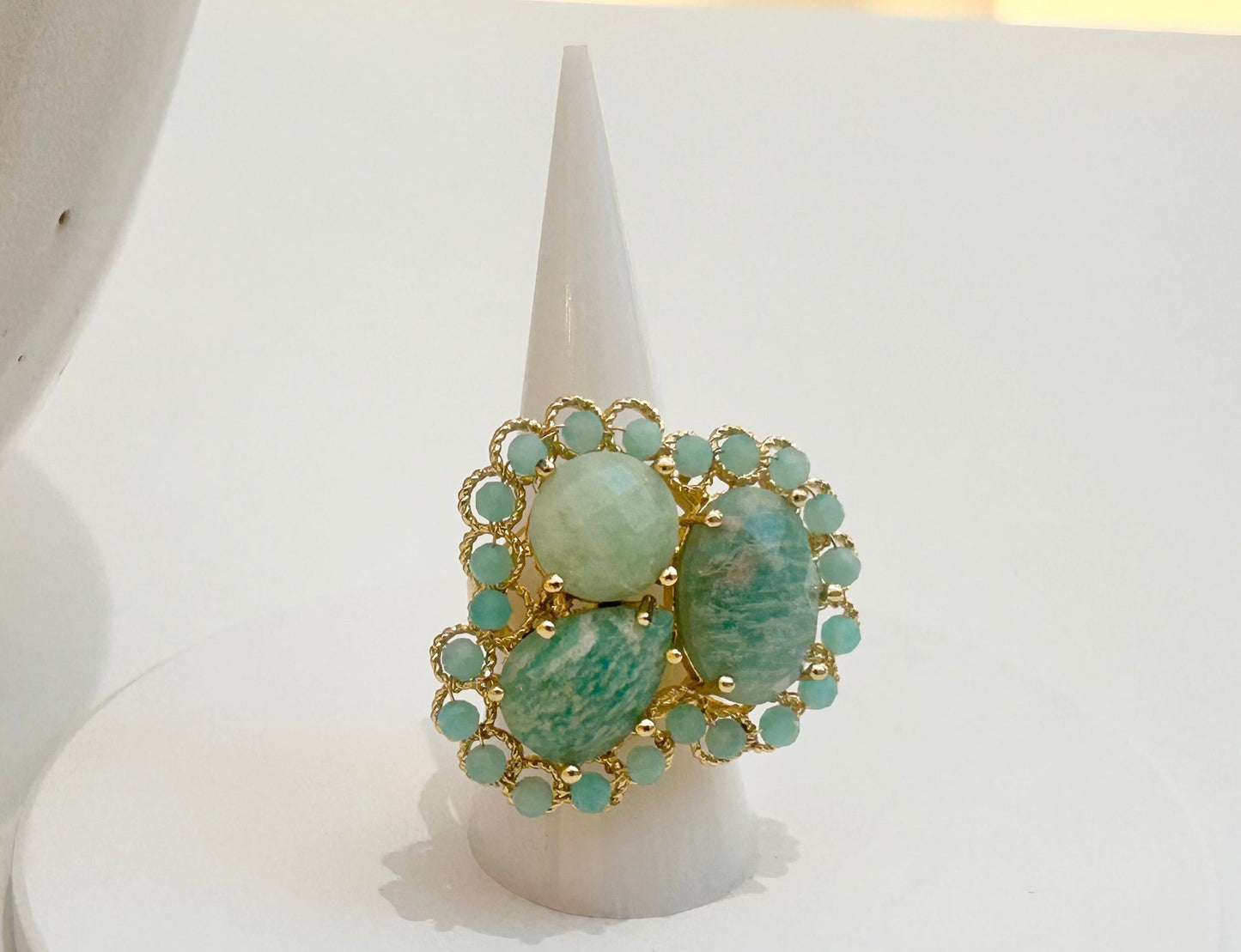 
                  
                    A gold ring with green marbled amazonite gemstones displayed on a white cone stand, set against a white surface with a neutral ceramic vessel in the background.
                  
                
